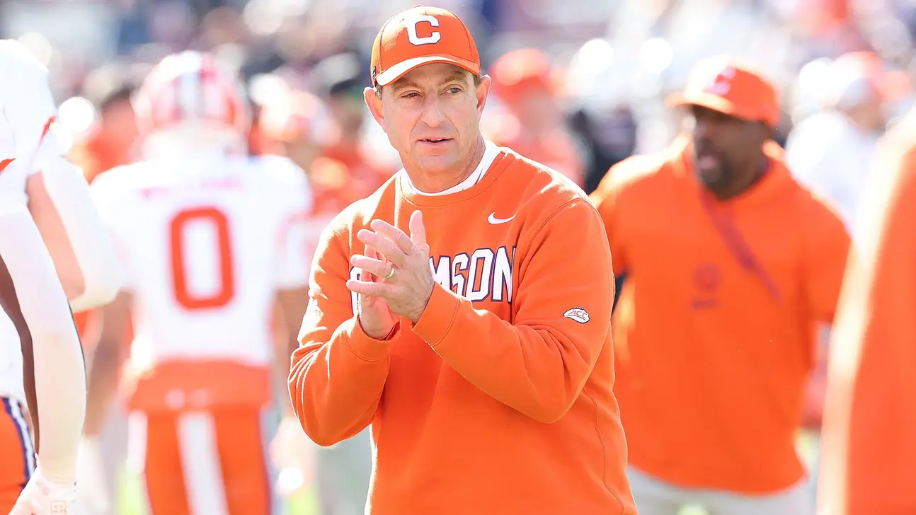 Clemson coach in orange sweatshirt and cap claps on football sideline during a game