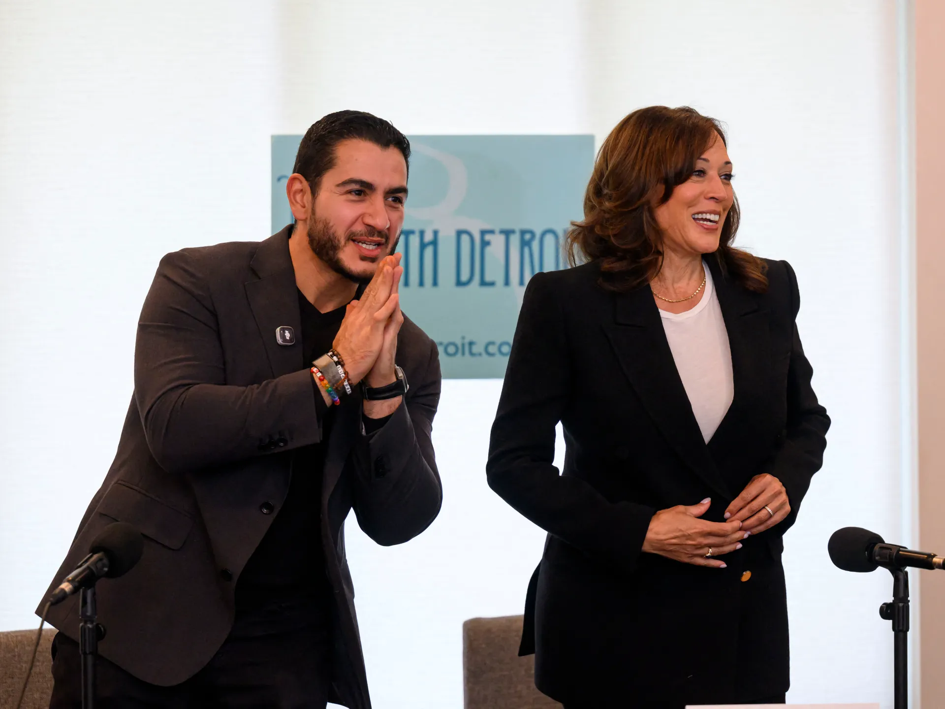 Two people in dark suits smile; one claps as the other stands beside them at microphones.