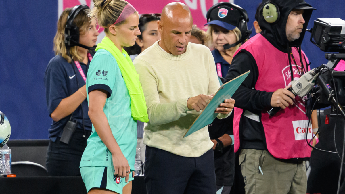 A soccer player and coach examine a tablet as camera crew members gather around them.
