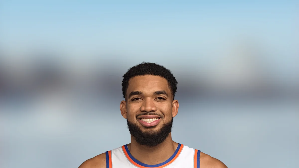 Smiling man with short curly hair and beard wearing a white basketball jersey with blue and orange trim