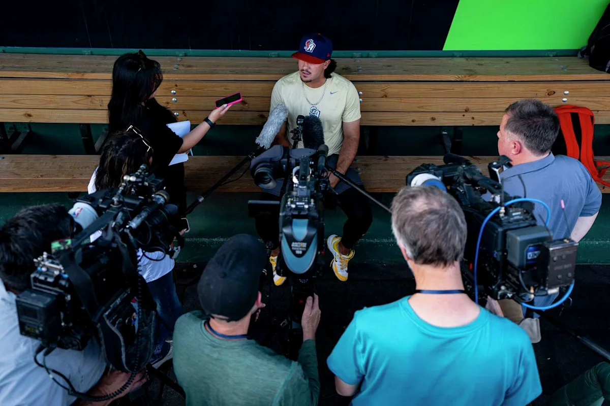 Baseball player seated in a dugout speaking with reporters and surrounded by television cameras
