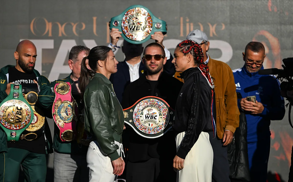 Two female boxers face each other while officials display multiple championship belts onstage