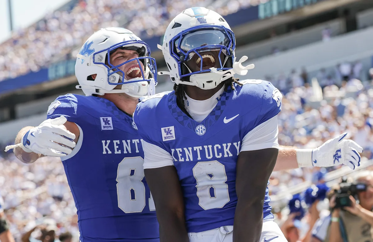 Two Kentucky football players in blue uniforms and white helmets celebrate together on the field.