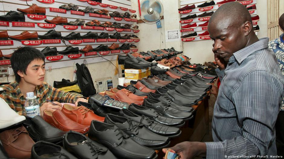 Customer examines leather shoes at a crowded shoe shop counter while a shopkeeper sits behind it.