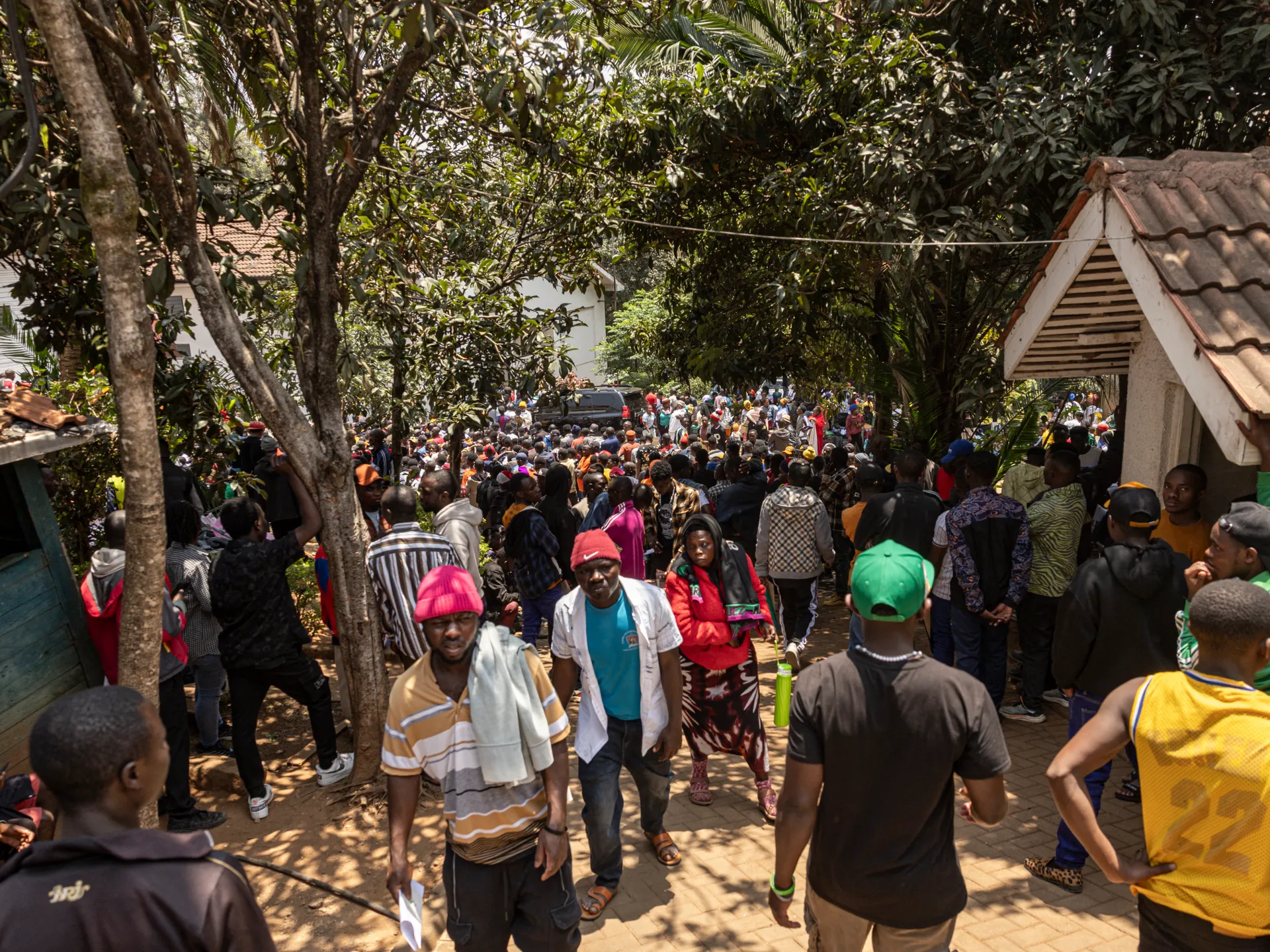 Large crowd gathered beneath trees outside a tiled-roof building, with people walking through a shaded courtyard.