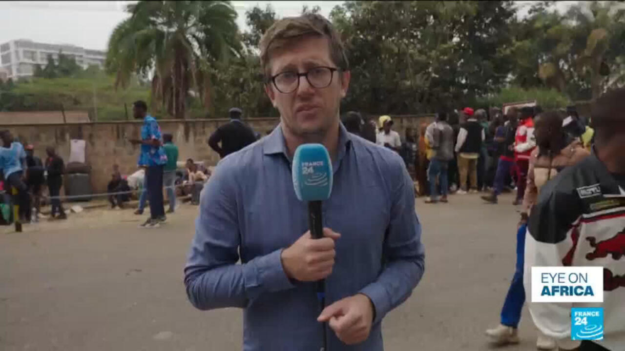 Reporter holding a France 24 microphone stands before a crowd near a wall and trees.