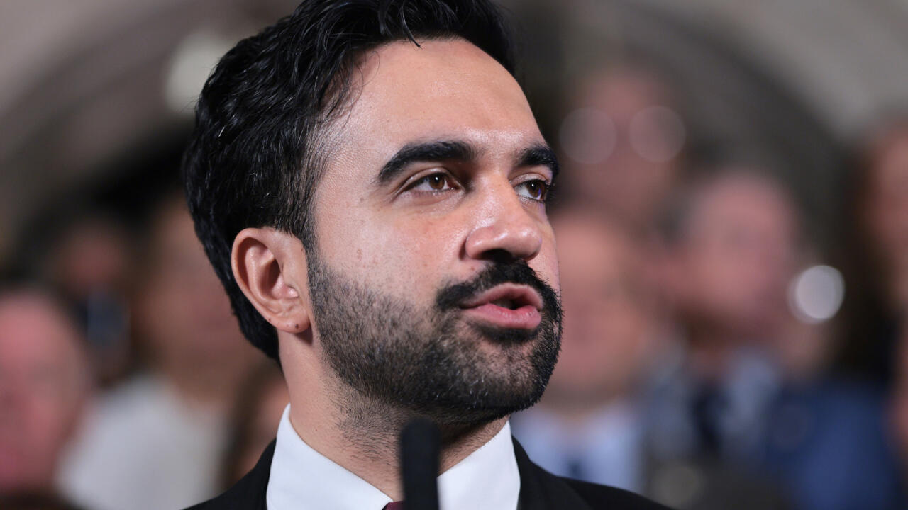 Bearded man in a dark suit speaks while surrounded by a blurred crowd.