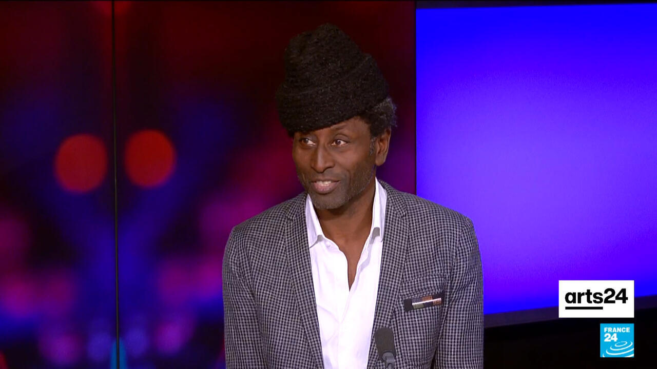 Man in patterned jacket and hat appears on a brightly lit televised arts program