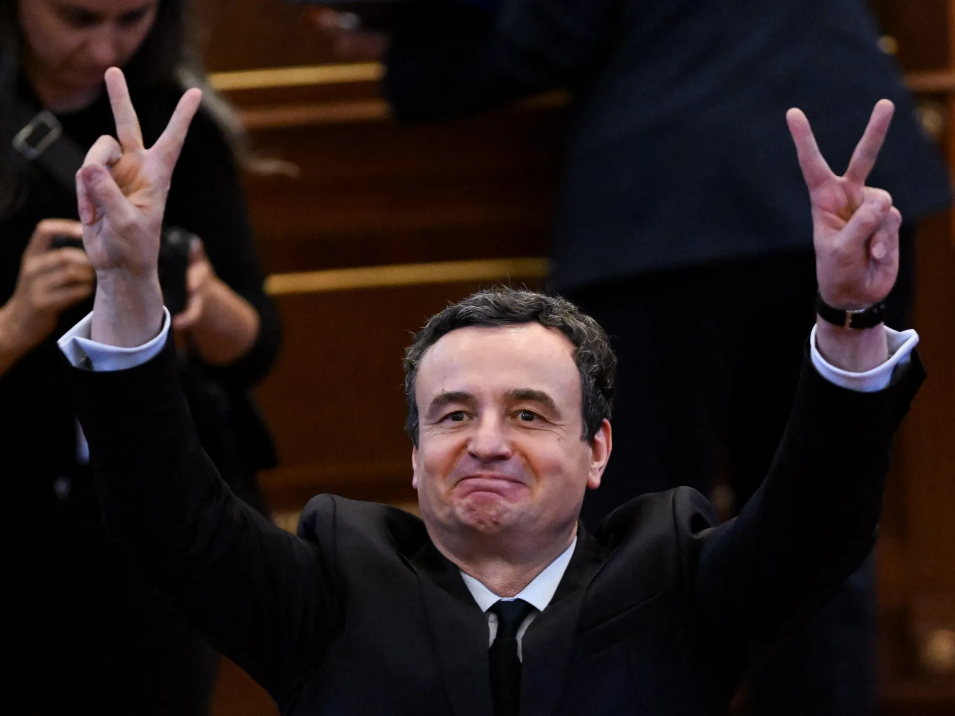 Man in dark suit raises both hands, making V signs, inside a formal chamber.