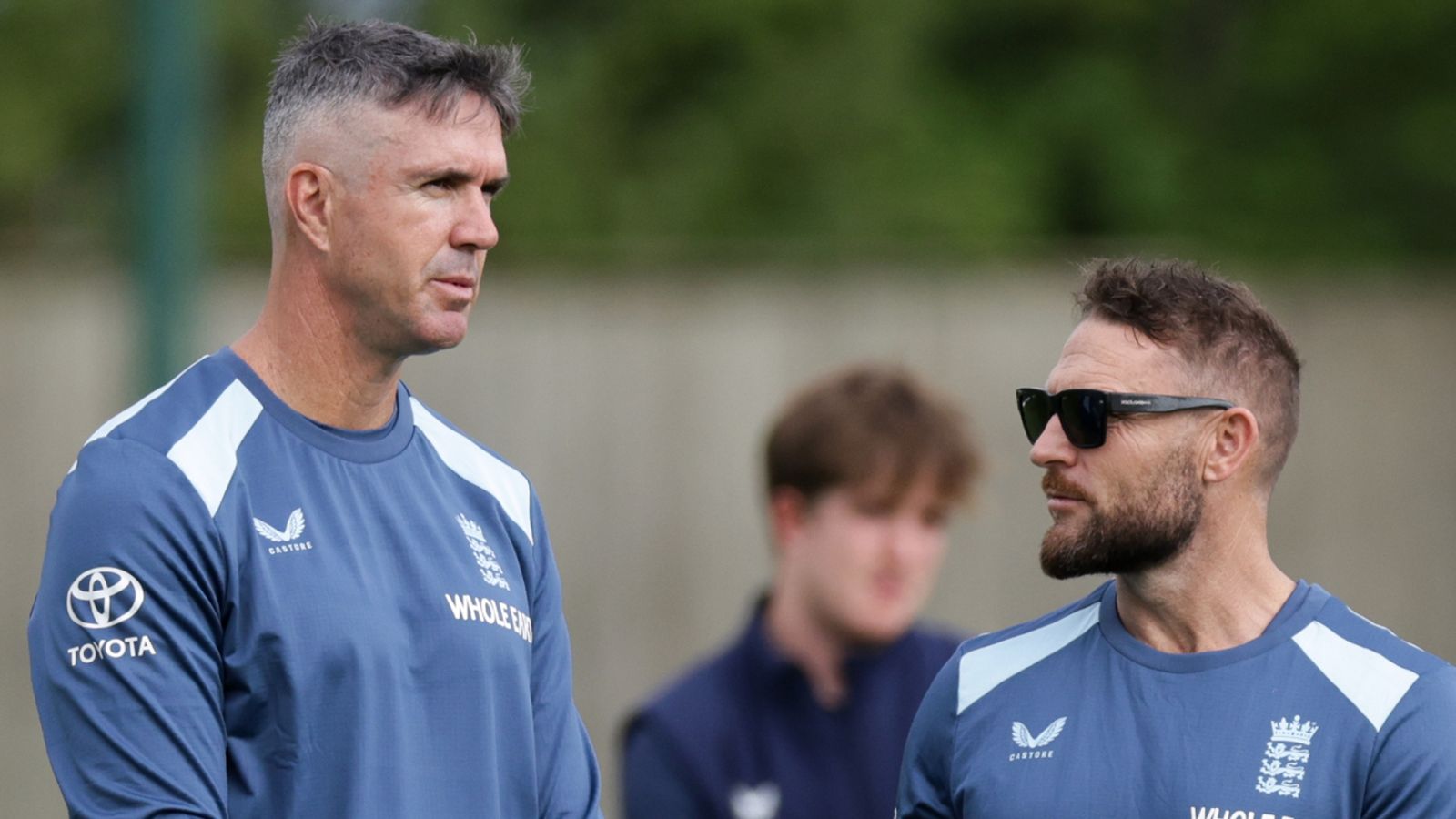 Two men in blue England training shirts talk outdoors, with a blurred person behind them.