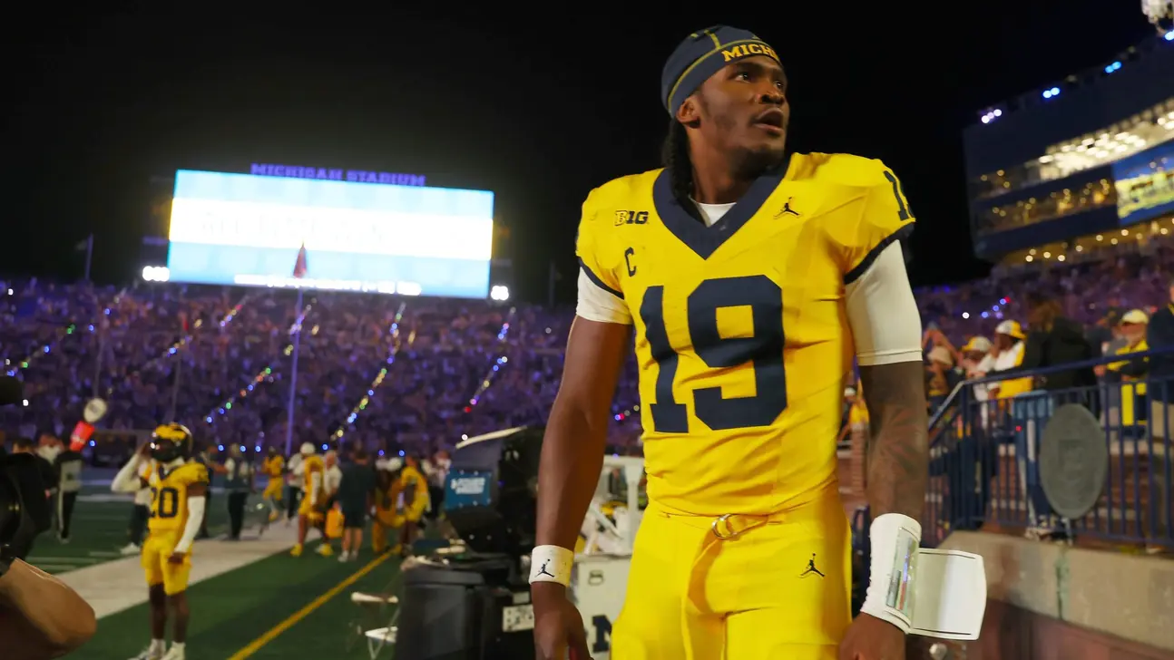 Football player in yellow Michigan uniform walking past the sideline during a nighttime game