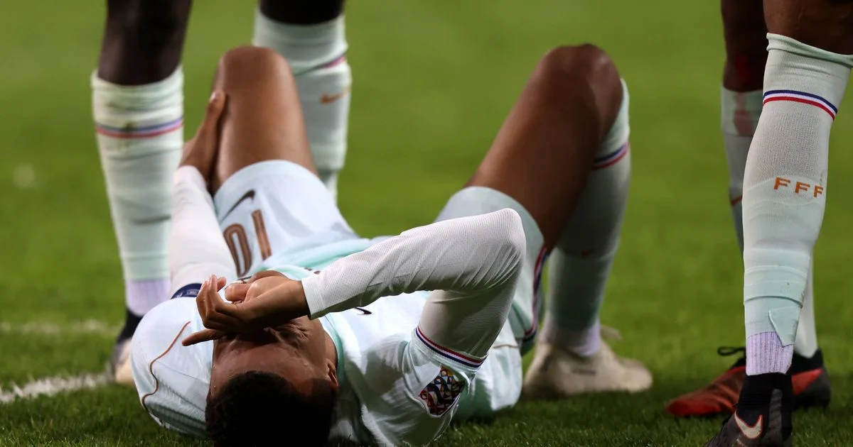 A footballer in a white kit lies on the grass with his hands over his face as teammates stand nearby.