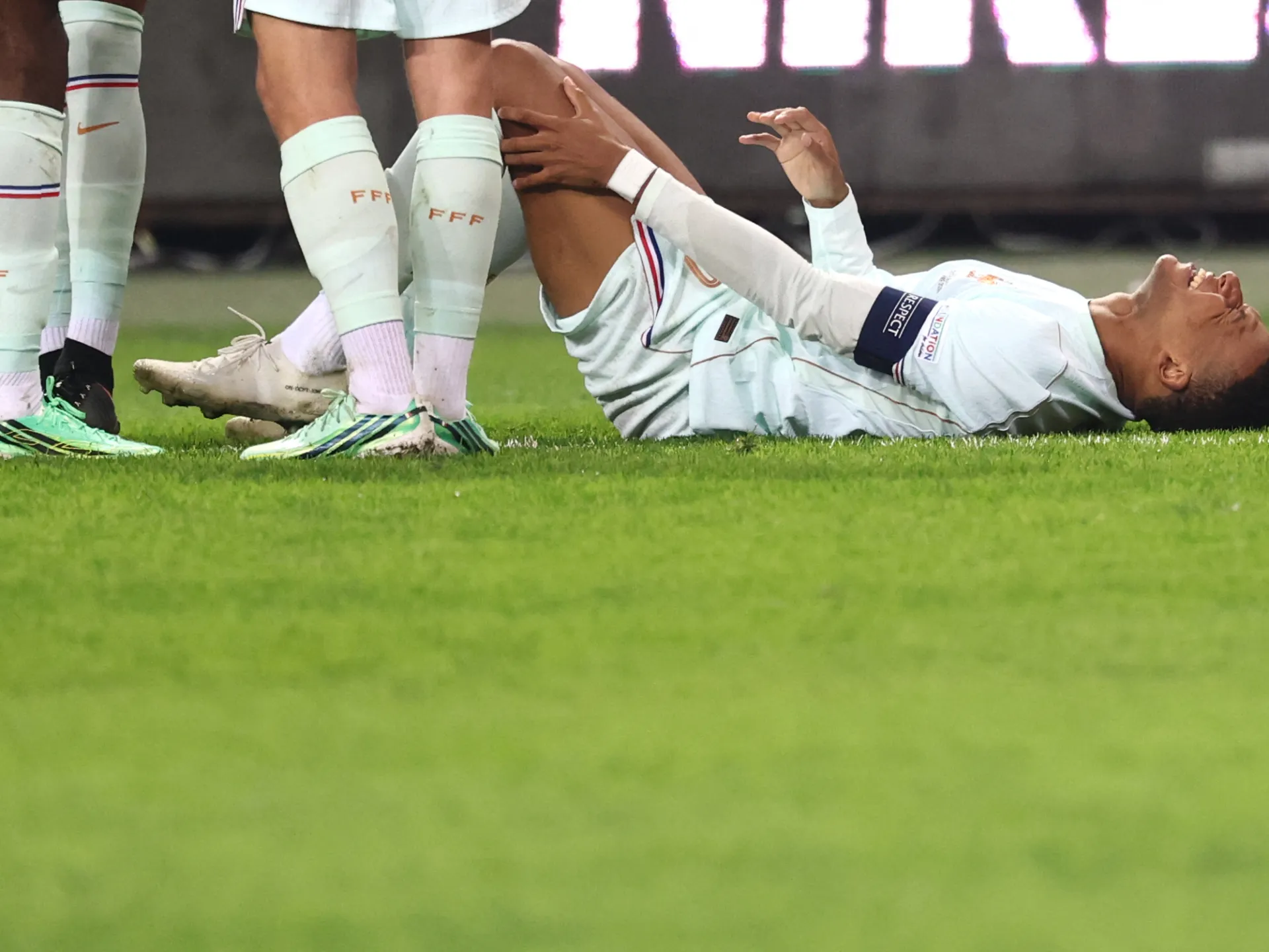 A footballer in a white kit lies on the pitch as teammates stand nearby.