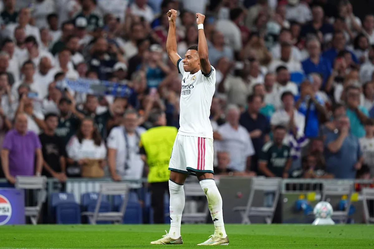 Footballer in white Real Madrid kit raises both fists before cheering supporters.