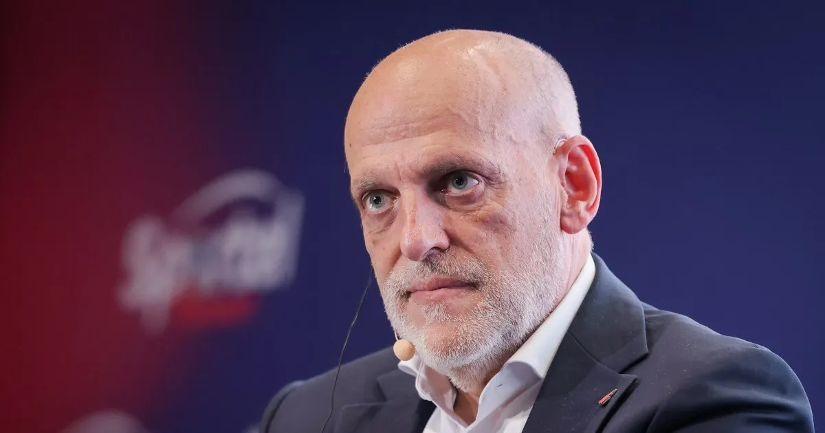 Bald man in suit and headset speaking against a dark blue and red backdrop