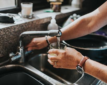 Person fills a drinking glass from a kitchen tap over a stainless-steel sink.
