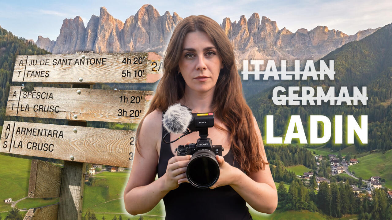 Woman holding a camera before Dolomite mountains, trail signs, and text reading Italian, German, Ladin.