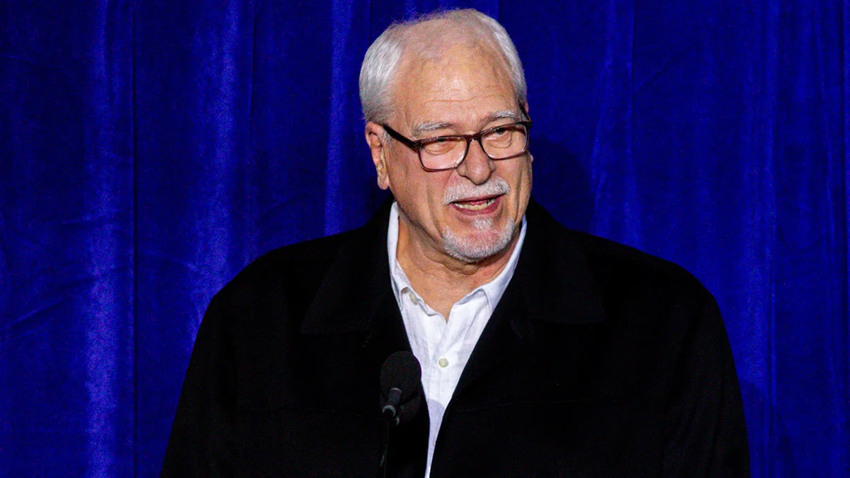 Gray-haired man wearing glasses and a black jacket speaks before a blue curtain.