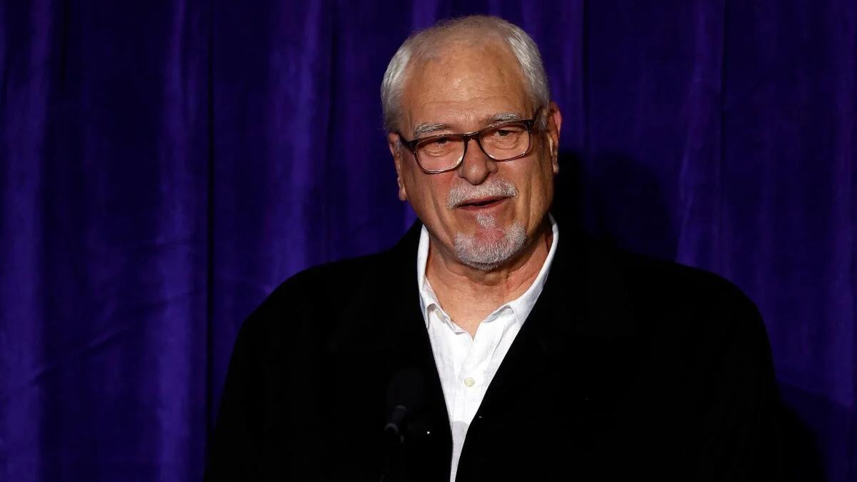 Older man with gray hair and glasses speaking at a microphone before a purple curtain