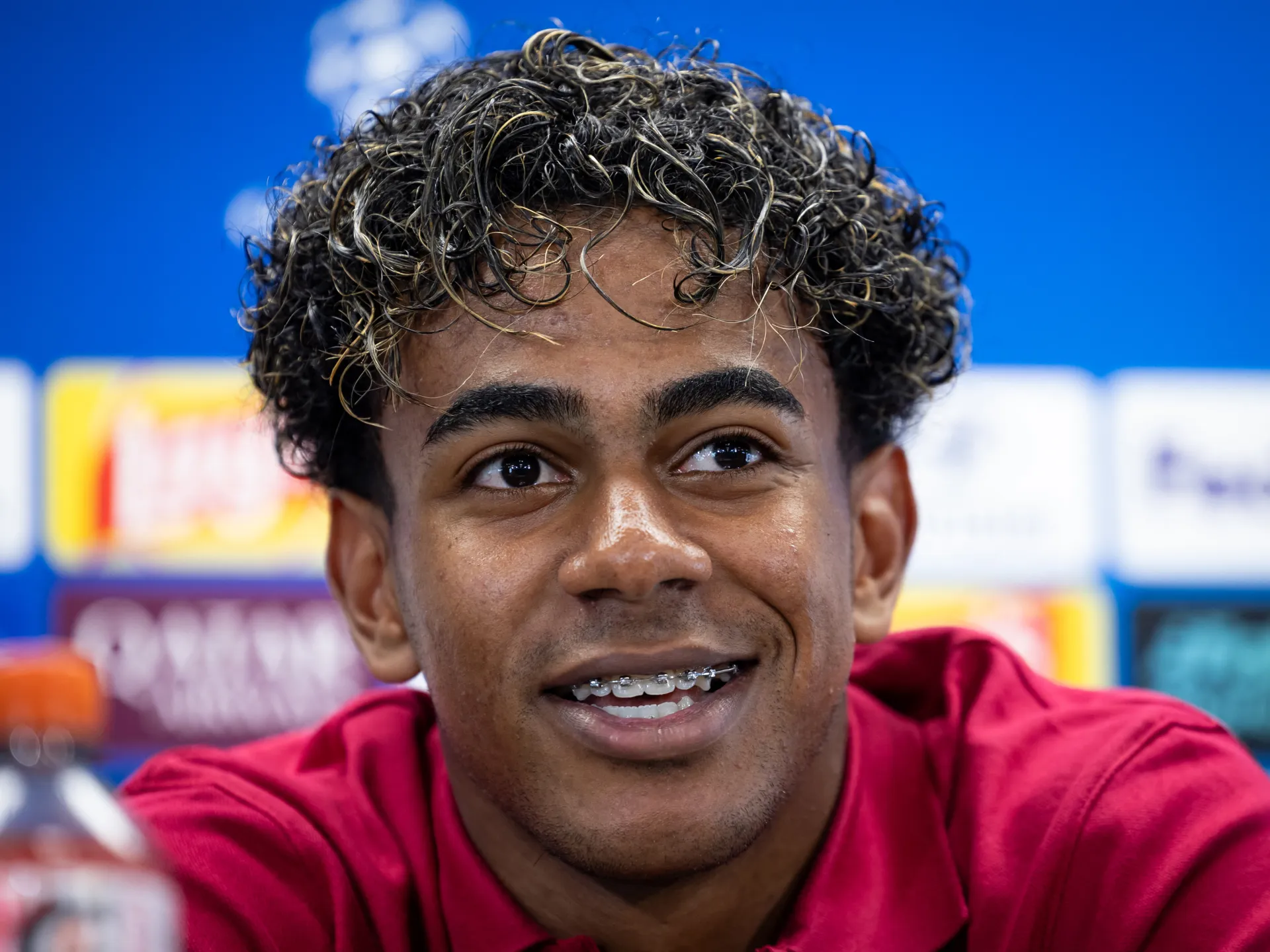 Close-up portrait of a smiling young footballer with curly hair and braces, wearing a red shirt.