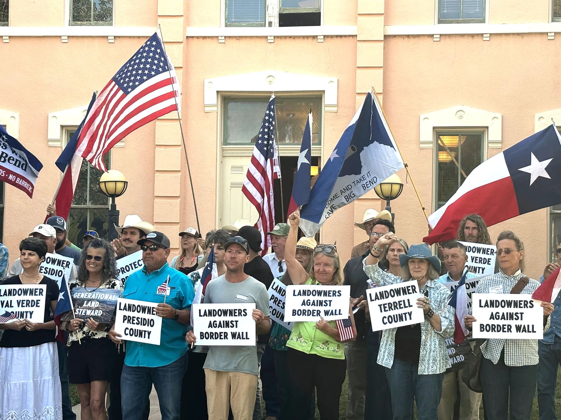 Group of people holding anti-border-wall signs and American and Texas flags outside a building