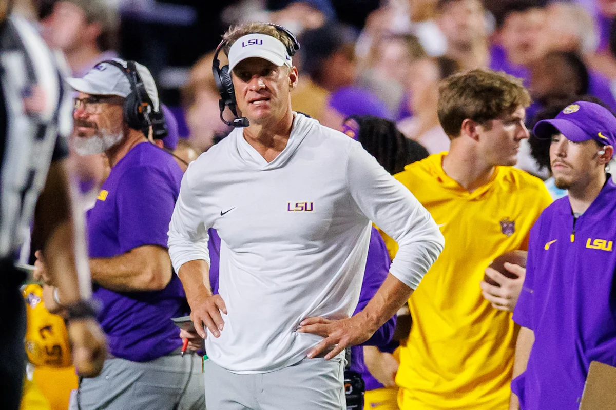 LSU coach in white visor and headset stands on sideline among players and staff.