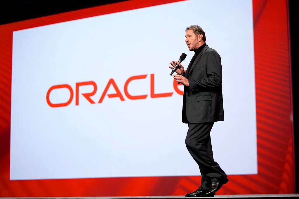 Man in black suit holding a microphone onstage before a large Oracle logo