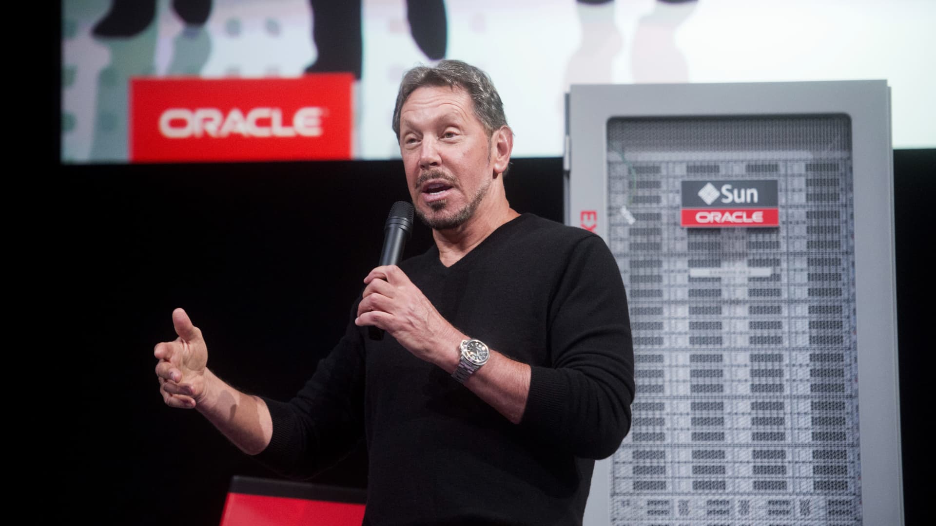 Man in black sweater speaks into a microphone beside an Oracle-branded server display.