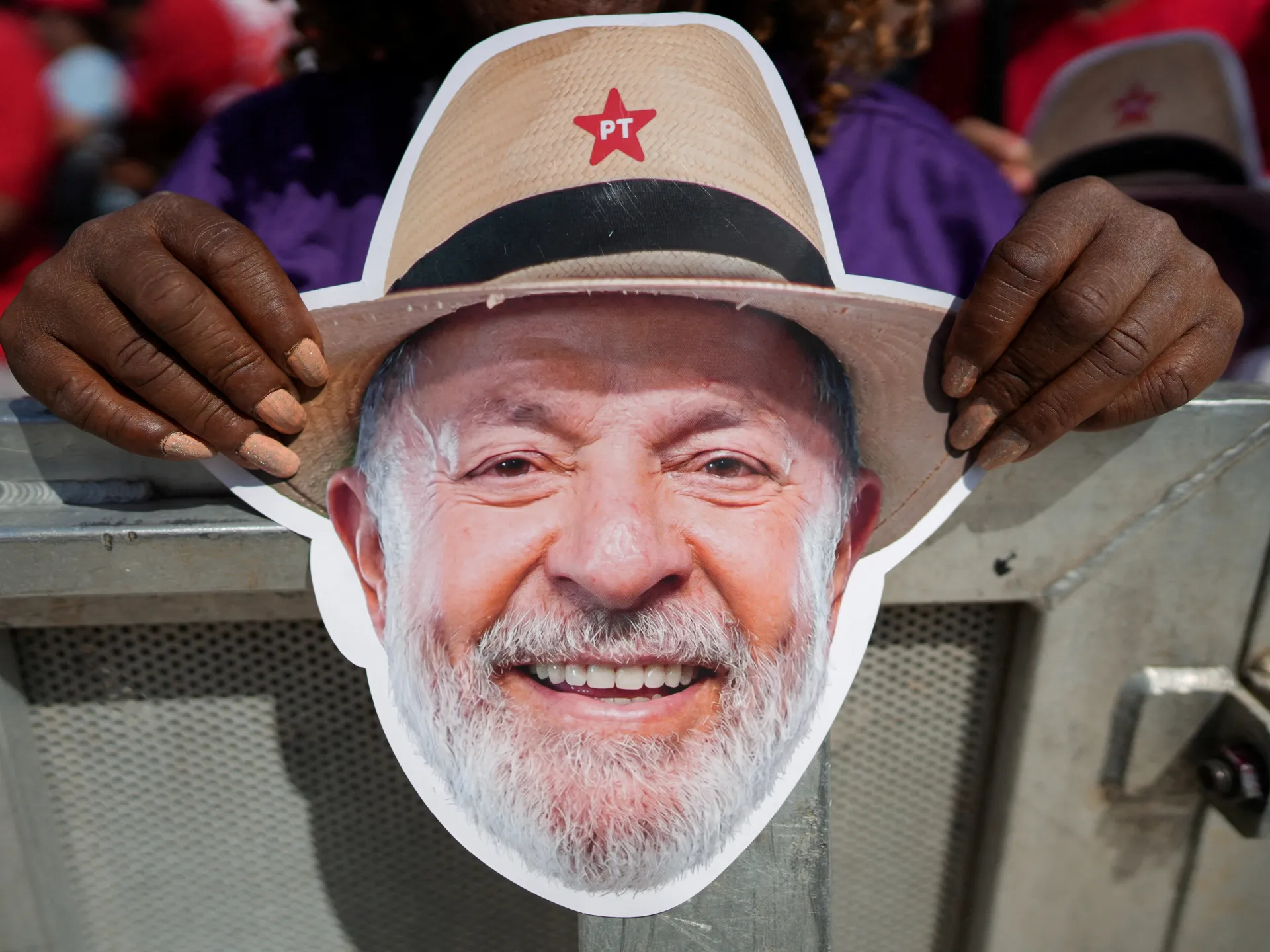 Smiling bearded man’s face printed on a political mask beneath a hat marked PT