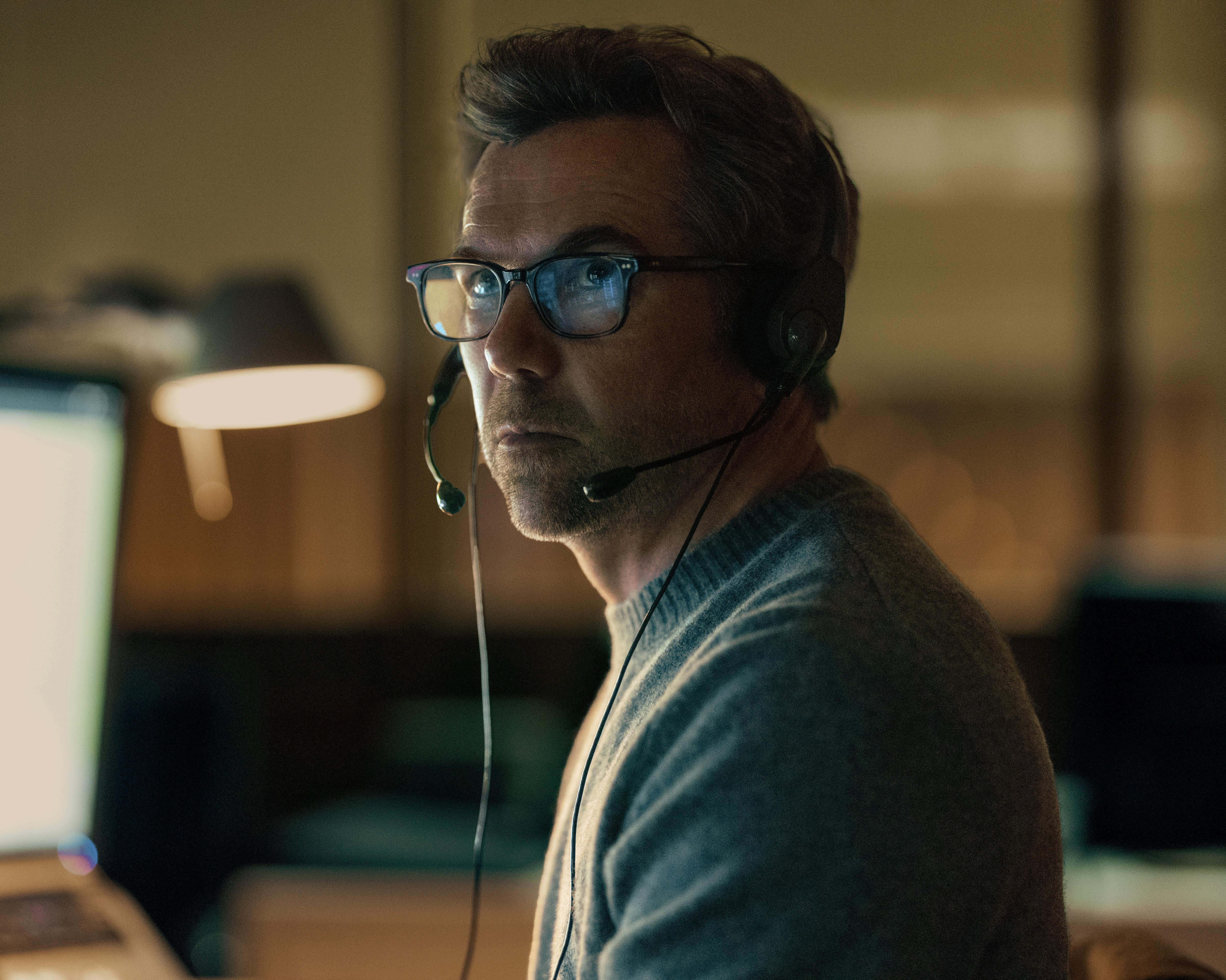 Man wearing glasses and a headset sits at a computer in a dimly lit room.