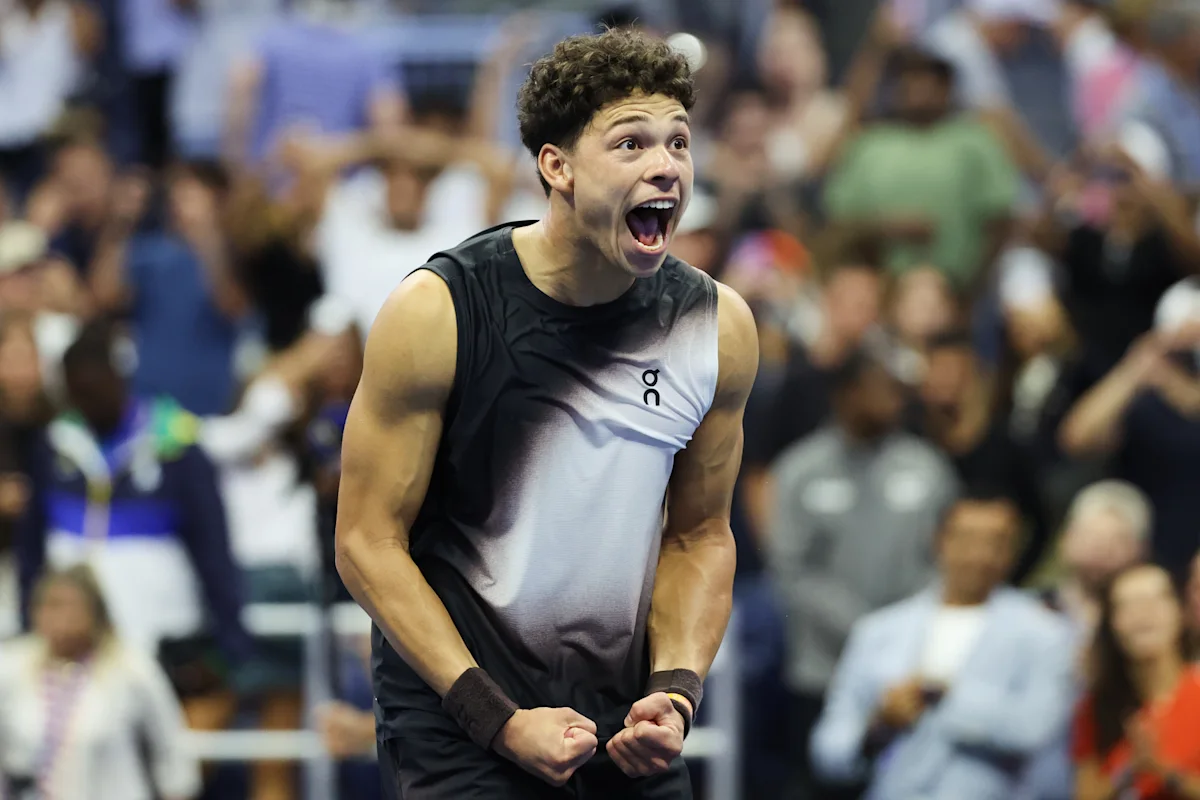 Tennis player in a sleeveless black-and-white shirt celebrates with clenched fists before a blurred crowd.
