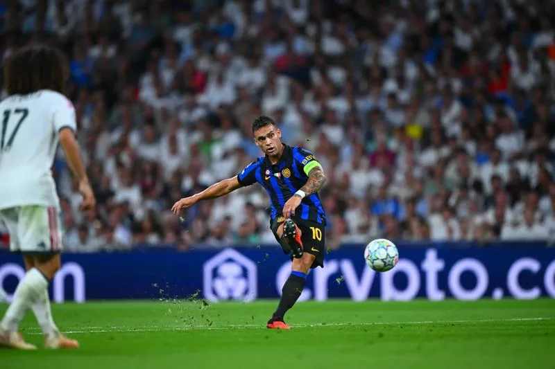 Footballer in blue-and-black kit strikes the ball during a match before a blurred crowd.