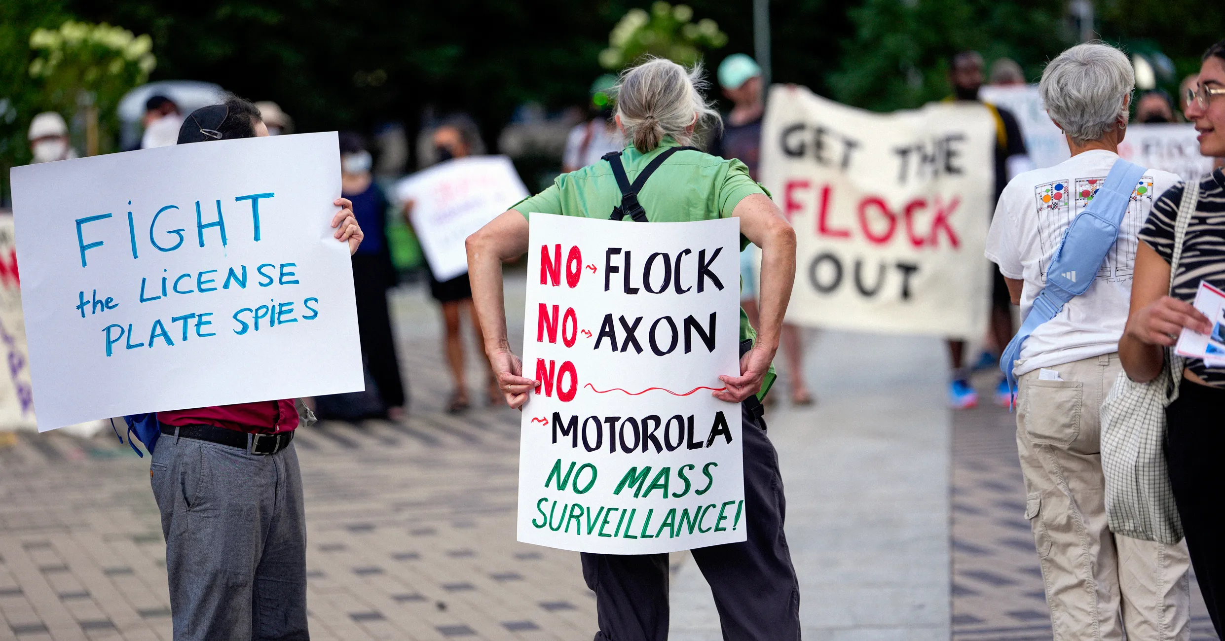 Protesters hold signs opposing Flock cameras, Axon, Motorola and mass surveillance.