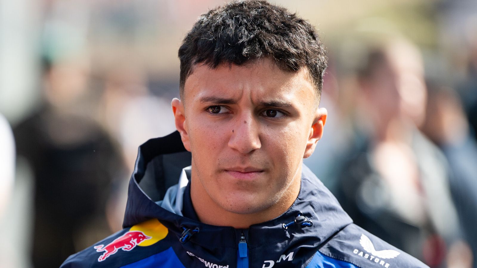 Young male driver in a Red Bull racing jacket looks toward the camera with a blurred crowd behind him