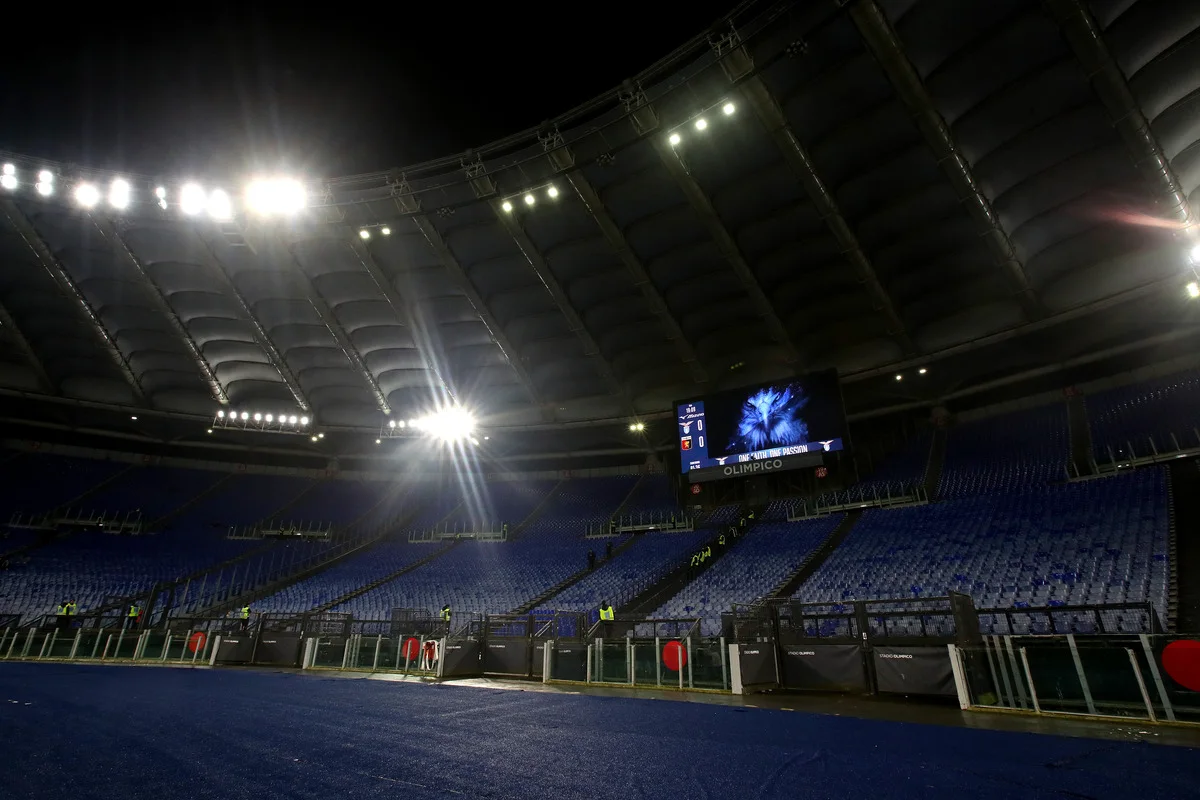 Empty blue-seated stadium under bright floodlights, with a scoreboard and dark running track.