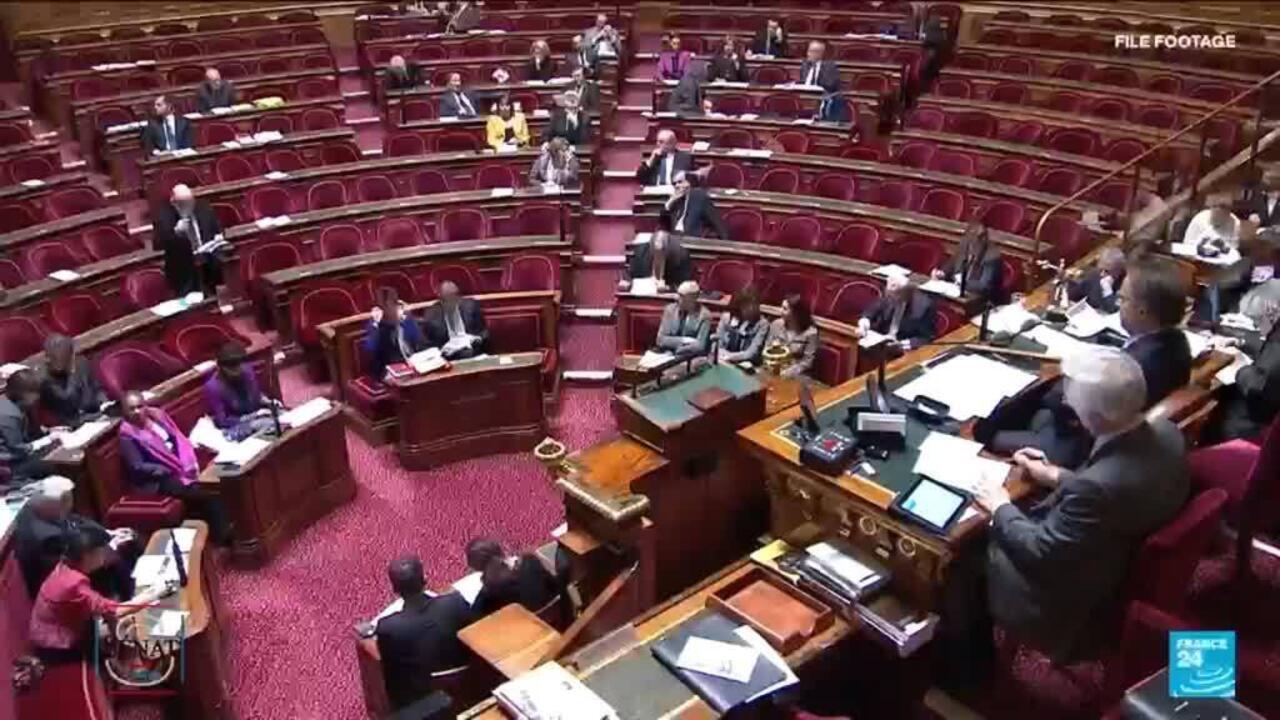Wide view of a sparsely attended parliamentary chamber with red seats, desks, and people gathered near the front.