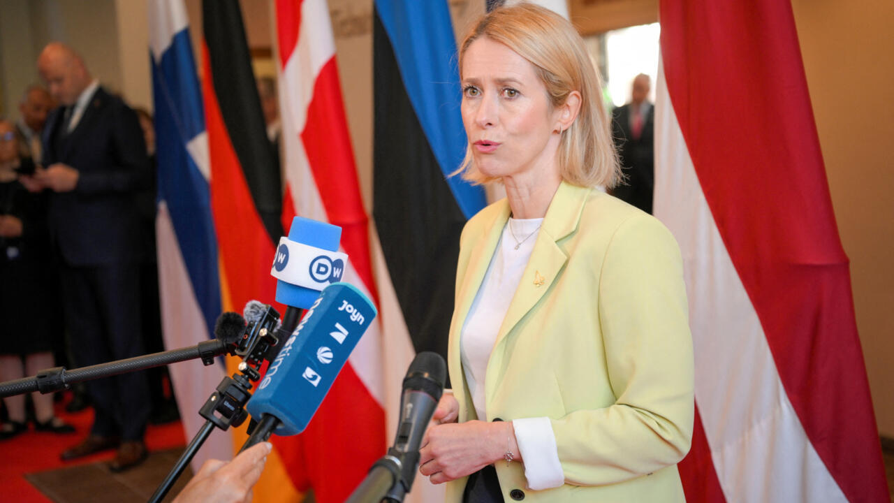 Blonde woman in pale yellow blazer speaks to reporters amid international flags and microphones