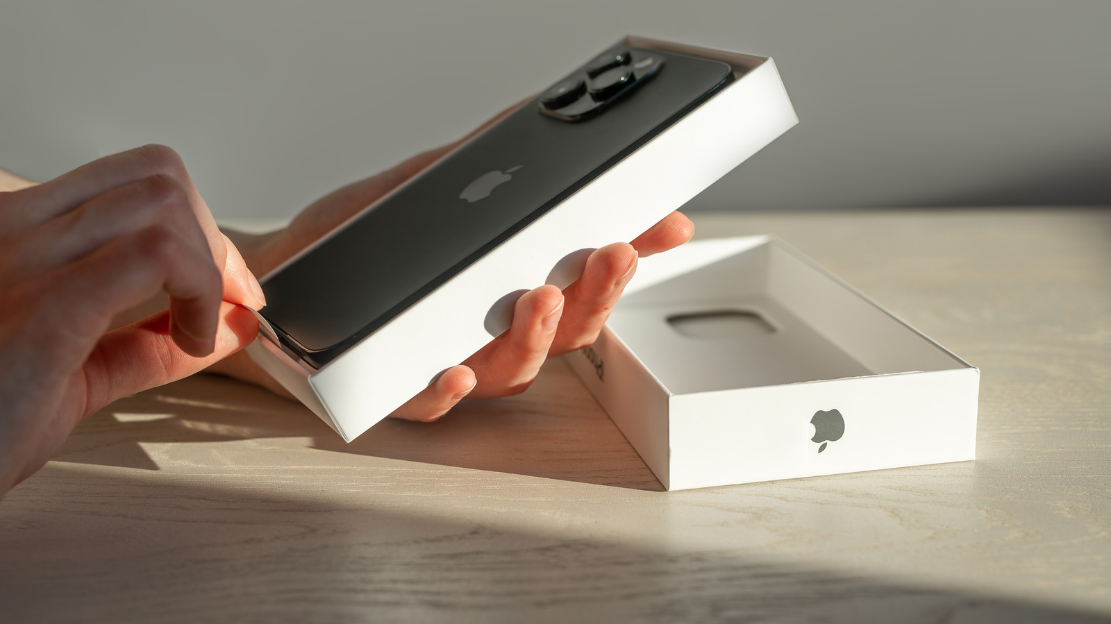 Hands lift a black iPhone from its white box on a sunlit wooden table.