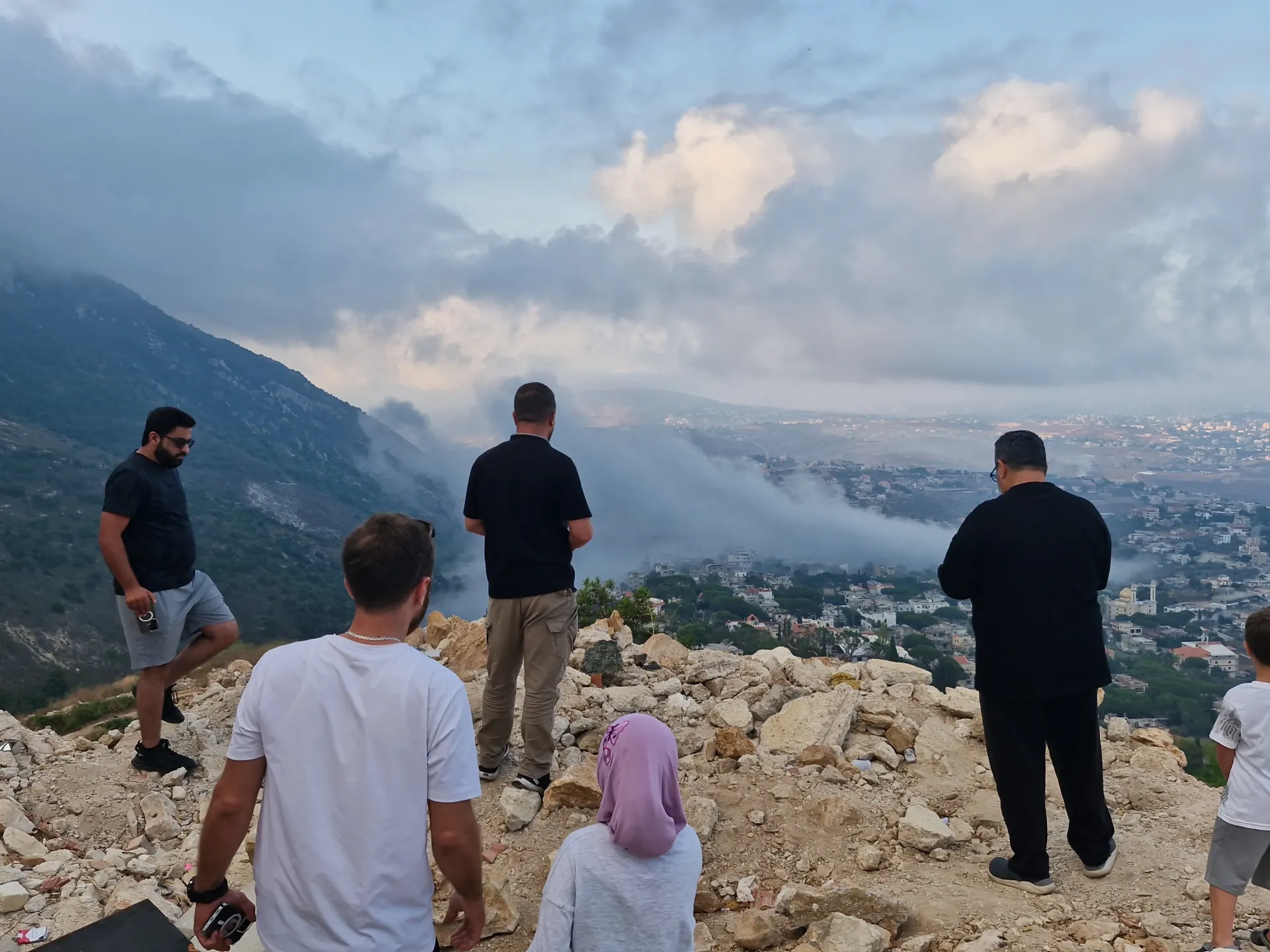 People stand on a rocky hillside overlooking a town partly covered by low clouds.