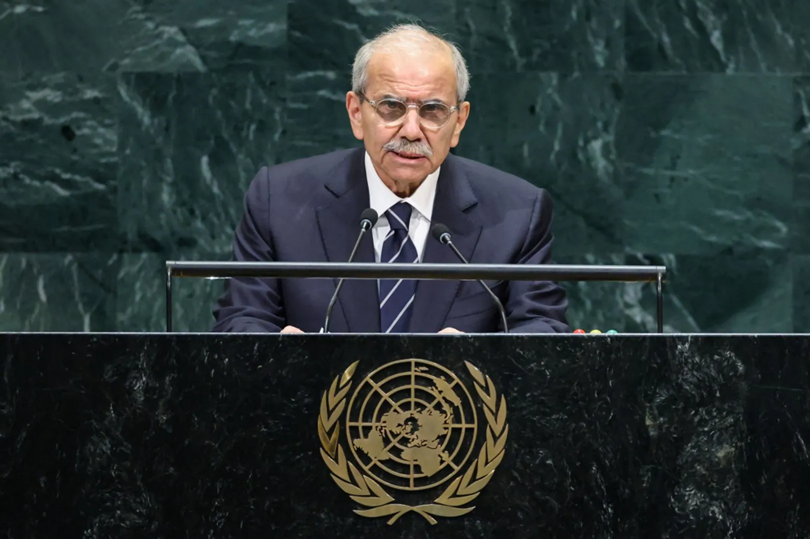 An older man in a dark suit and glasses speaks at a lectern before a green marble wall.