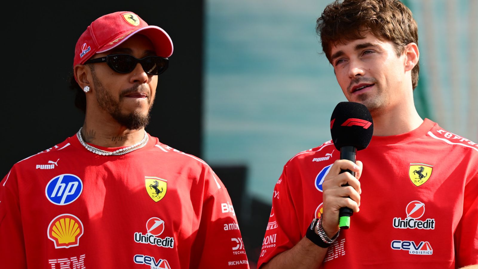 Two Ferrari drivers in red team shirts, one wearing sunglasses and the other holding a microphone