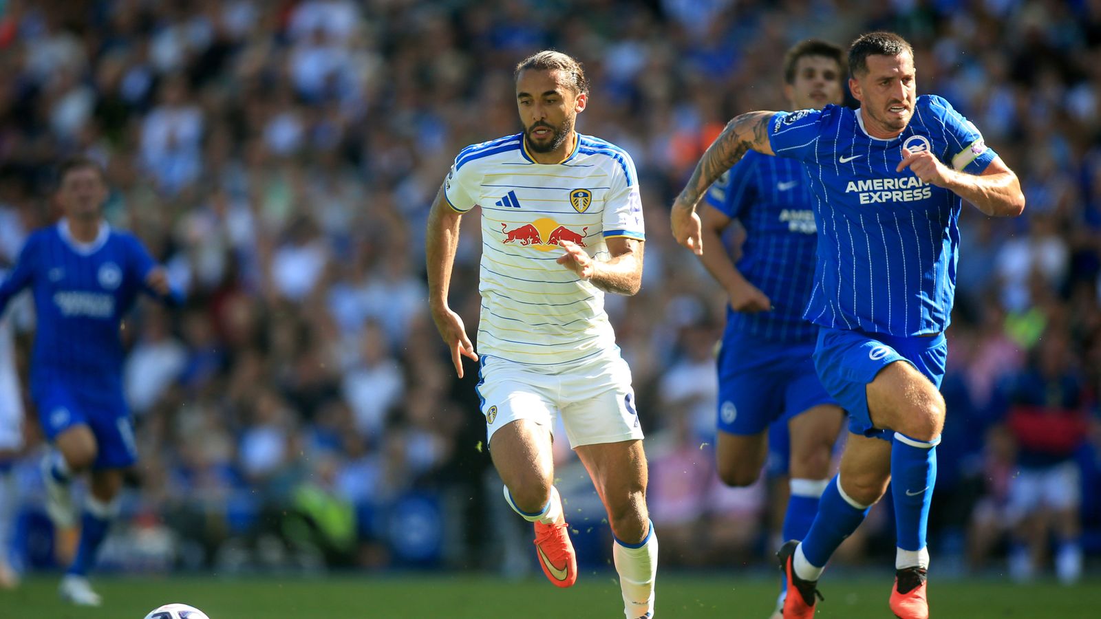 Two footballers in Leeds and Brighton kits sprint during a match, with a blurred crowd behind them.