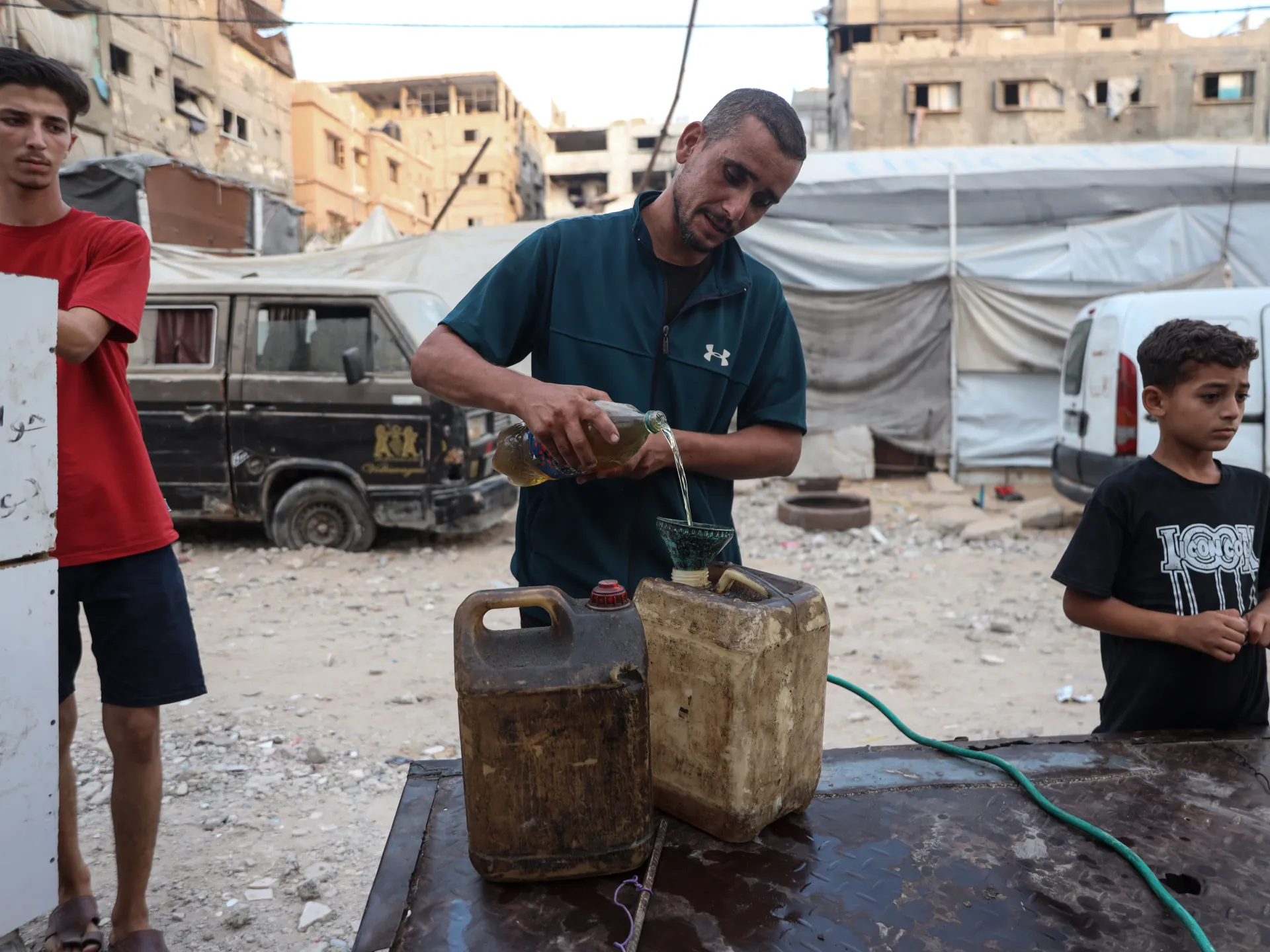 Man pours liquid through a funnel into a jerrycan as two boys stand nearby among damaged buildings.