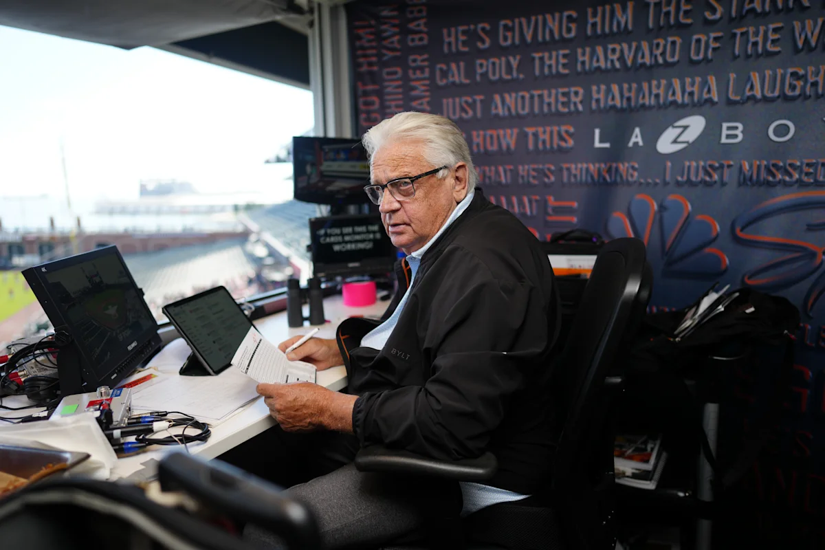 Gray-haired man seated at a broadcast desk, holding papers beside monitors in a stadium booth.