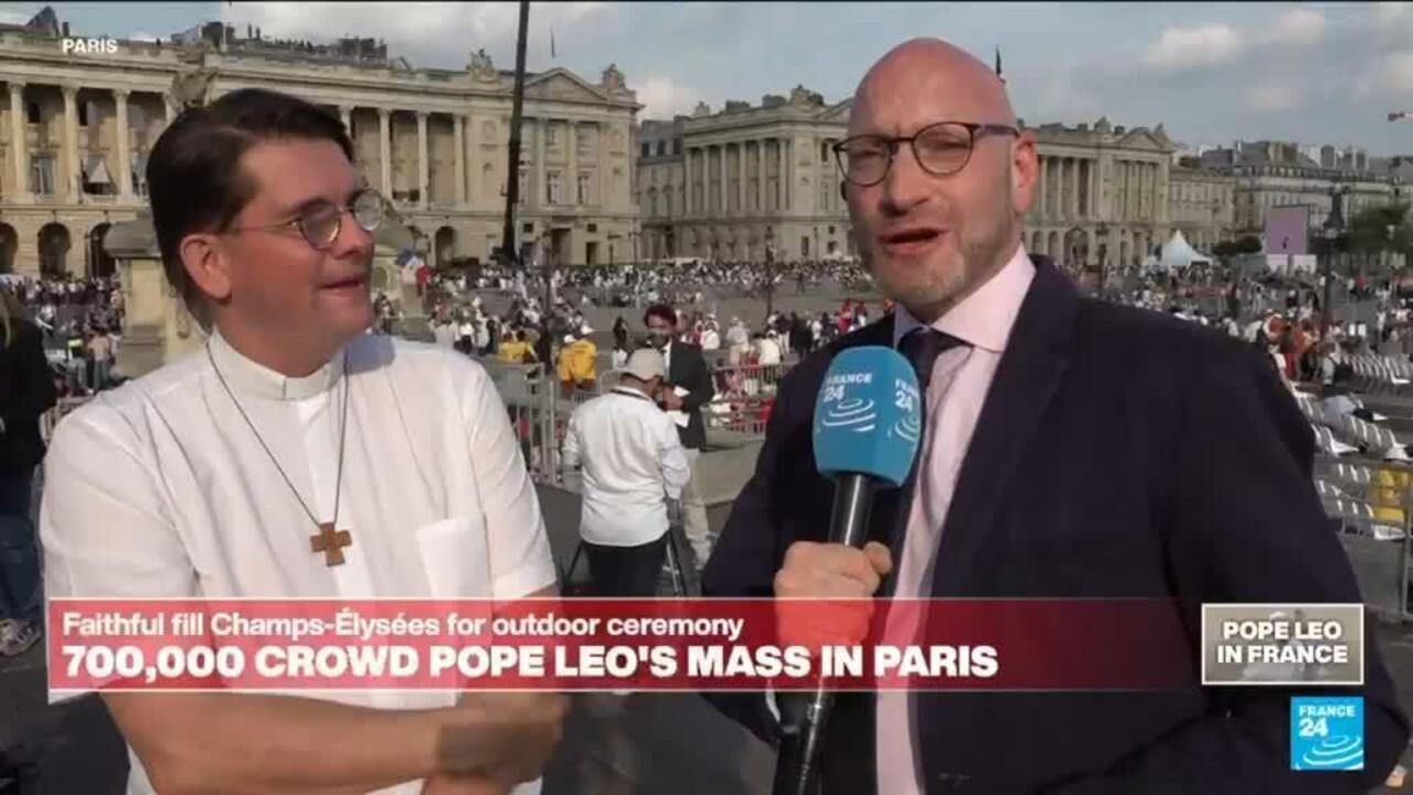 A France 24 reporter interviews a man in clerical clothing before a large outdoor crowd and historic buildings.