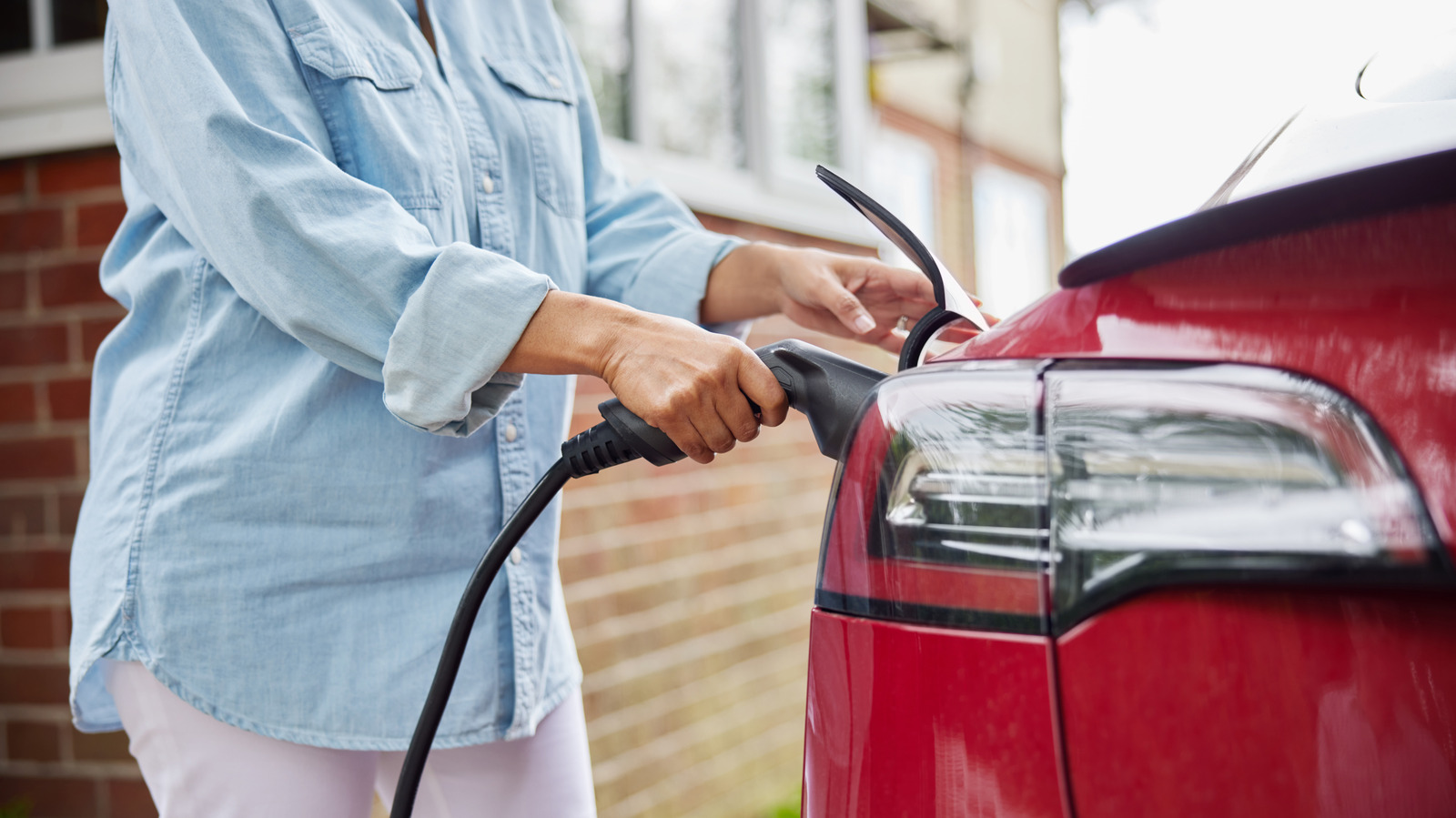 Person plugging a charging cable into the rear of a red electric car outside a brick building.
