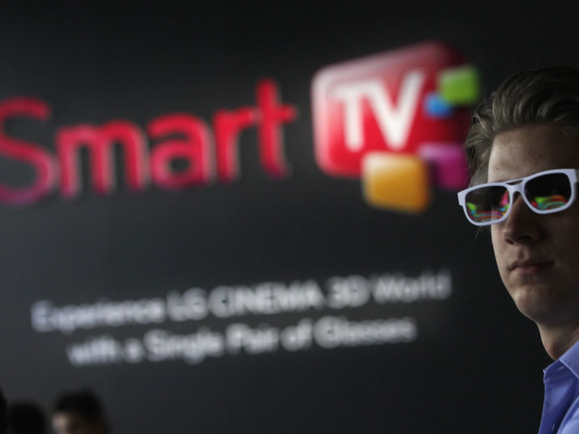 Man wearing white glasses stands beside a blurred Smart TV presentation screen