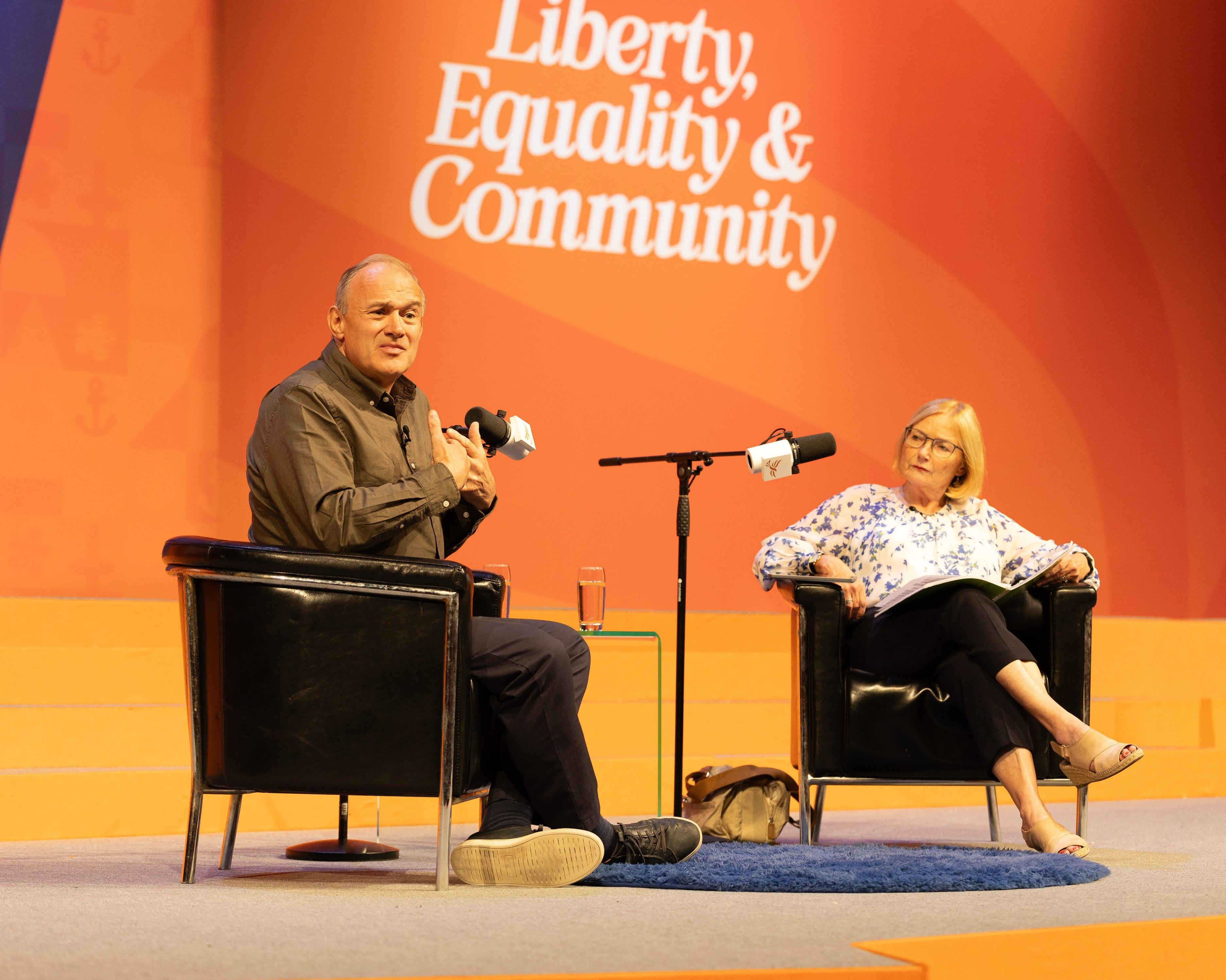 Two speakers sit onstage beside microphones against an orange backdrop reading “Liberty, Equality & Community”.