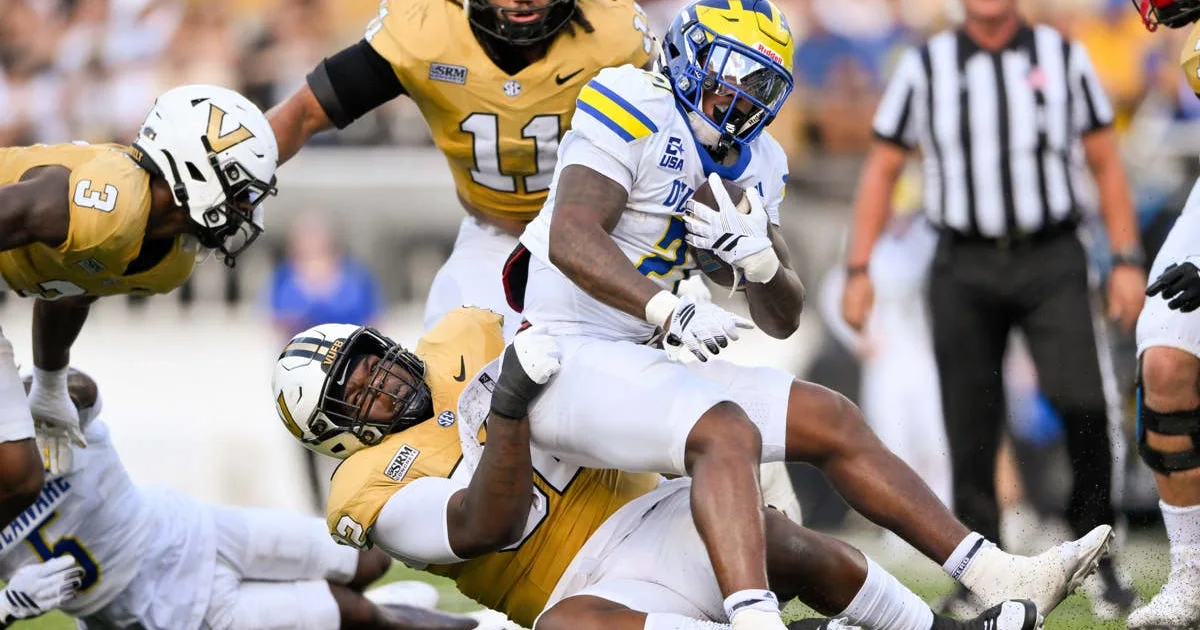 A Delaware player in white carries the ball as Liberty players in gold tackle him.