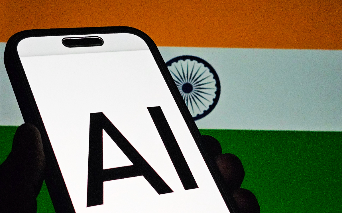 Illustration of a hand holding a smartphone displaying the letters AI, with an Ashoka Chakra behind it.